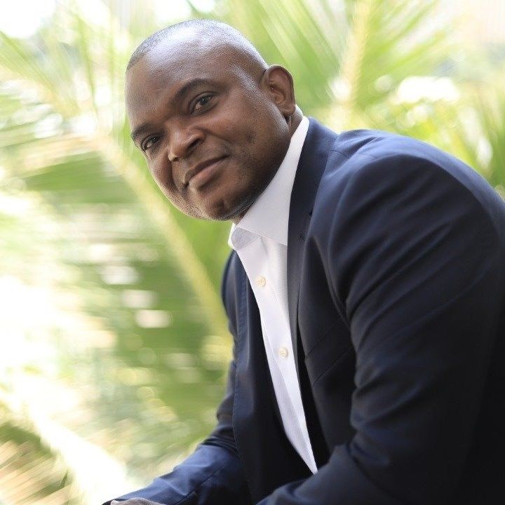Person wearing a dark suit and white shirt in an outdoor setting with green foliage in the background.
