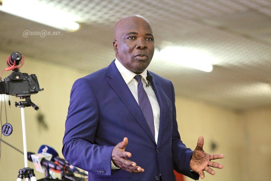 Man in a blue suit speaking with hand gestures, standing next to video camera equipment.